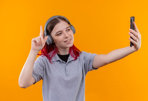 Sorridente giovane bella ragazza che indossa la maglietta grigia in cuffie punta il dito verso l'alto prendendo selfie su sfondo giallo isolato