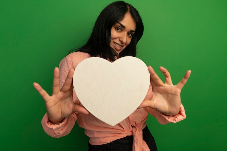 Foto gratuito sorridente giovane bella ragazza che indossa la maglietta rosa che tiene la scatola di forma del cuore isolata sul verde