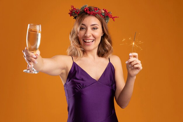Sorridente giovane bella ragazza che indossa un abito viola con ghirlanda che tiene le stelle filanti e porge un bicchiere di champagne alla telecamera isolata su sfondo marrone