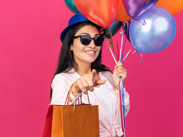 Sorridente giovane bella ragazza che indossa un cappello da festa che tiene palloncini con sacchetti regalo che ti mostrano un gesto