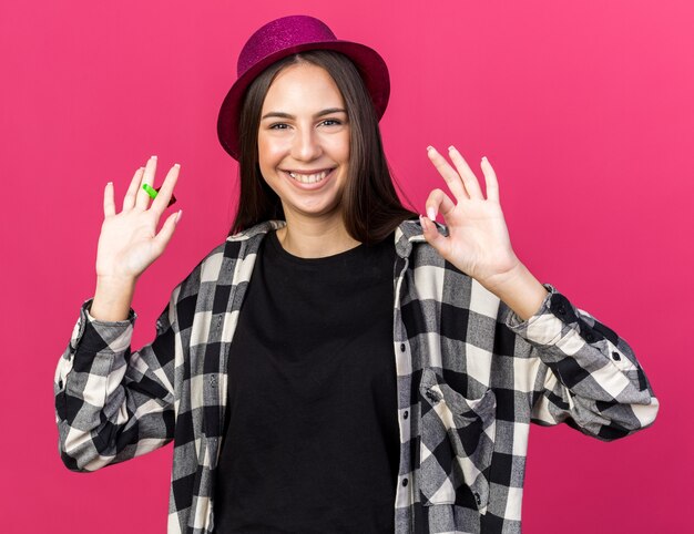 Sorridente giovane bella ragazza che indossa un cappello da festa che tiene un fischio di festa che mostra un gesto ok