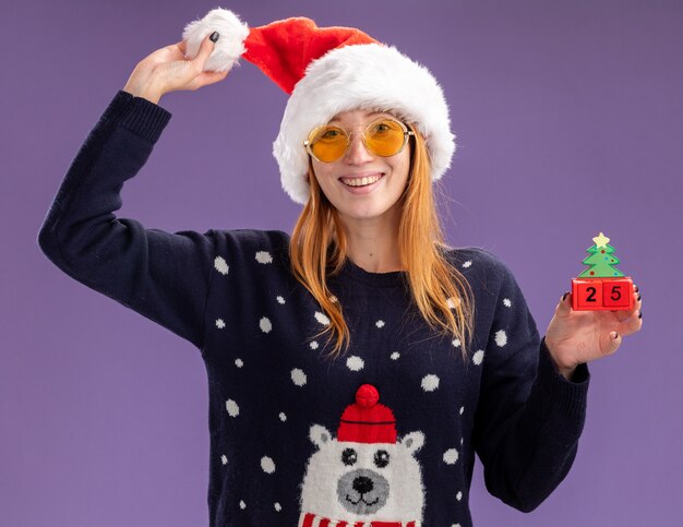 Sorridente giovane bella ragazza che indossa un maglione di natale e cappello con gli occhiali che tengono il giocattolo di natale e cappello isolato su parete viola