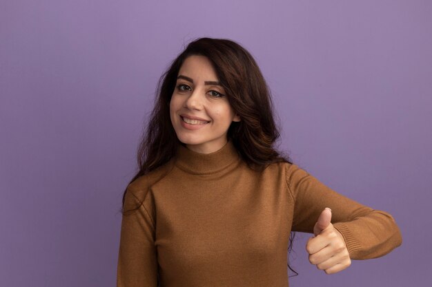 Sorridente giovane bella ragazza che indossa un maglione dolcevita marrone che mostra il pollice in su isolato sulla parete viola