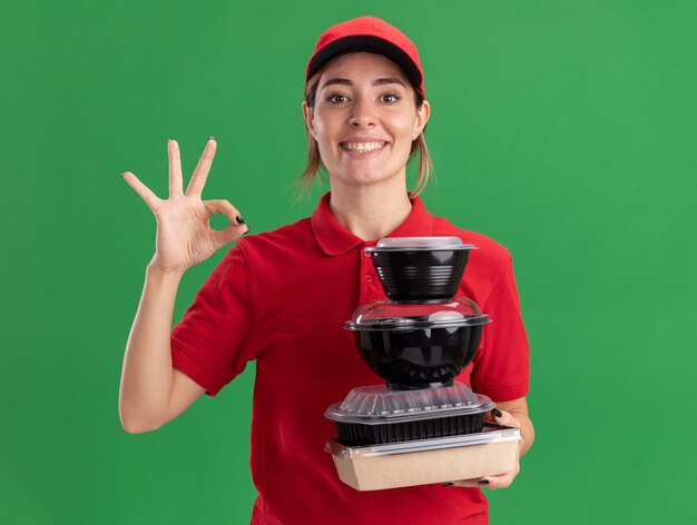 Sorridente giovane bella ragazza di consegna in gesti uniformi segno ok con la mano e tiene i contenitori per alimenti sul pacchetto alimentare sul verde