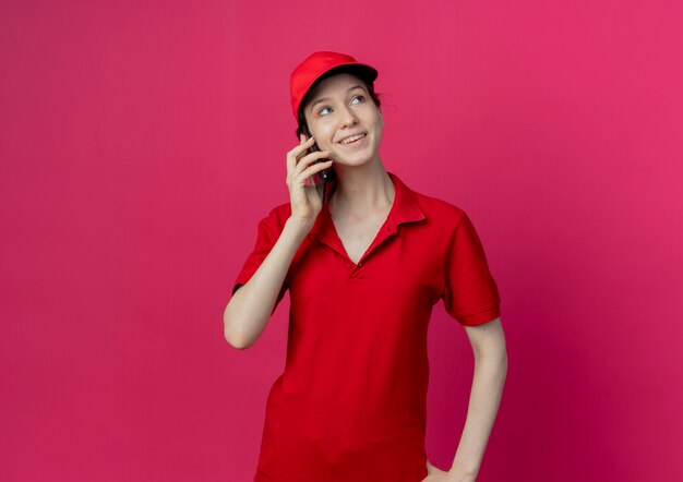 Sorridente giovane bella ragazza di consegna in uniforme rossa e cappuccio parlando al telefono guardando il lato isolato su sfondo cremisi con lo spazio della copia