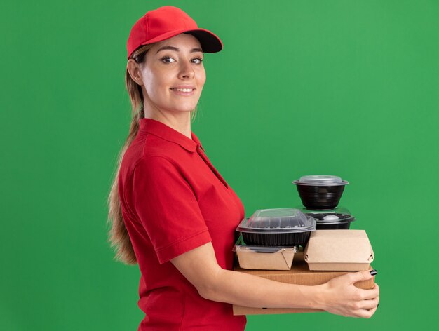 Sorridente giovane bella ragazza di consegna in uniforme si leva in piedi lateralmente tenendo i pacchetti di carta alimentare