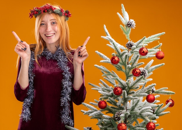 Sorridente giovane bella ragazza in piedi vicino albero di natale che indossa abito rosso e corona con ghirlanda sul collo punta verso l'alto isolato su parete arancione orange