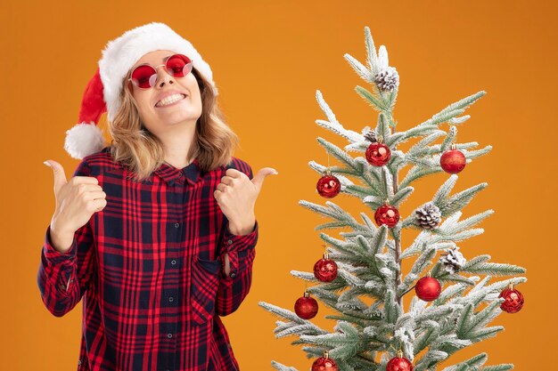 Sorridente giovane bella ragazza in piedi vicino albero di natale che indossa cappello di natale con gli occhiali che mostra sì gesto isolato su sfondo arancione