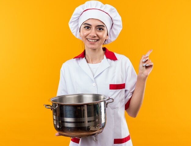 Sorridente giovane bella ragazza in uniforme da chef tenendo e guardando i punti di pentola a lato