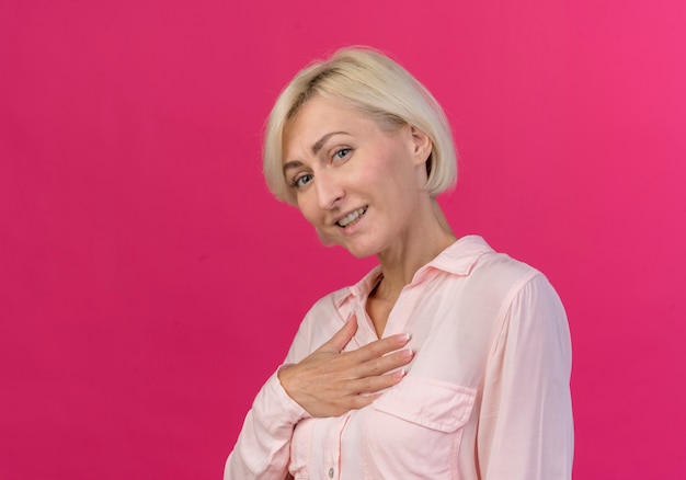 Sorridente giovane bionda donna slava in piedi in vista di profilo mettendo la mano sul petto guardando la telecamera isolata su sfondo rosa con spazio di copia