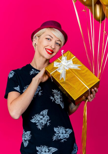 Sorridente giovane bionda festa donna che indossa il cappello del partito che tiene il contenitore di regalo e palloncini guardando davanti isolato sulla parete rosa