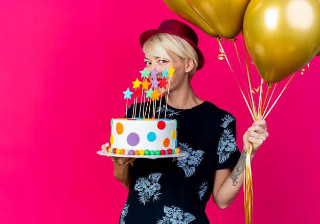 Sorridente giovane bionda festa donna che indossa il cappello del partito che tiene palloncini e torta di compleanno con le stelle guardando la parte anteriore isolata sulla parete rosa con spazio di copia
