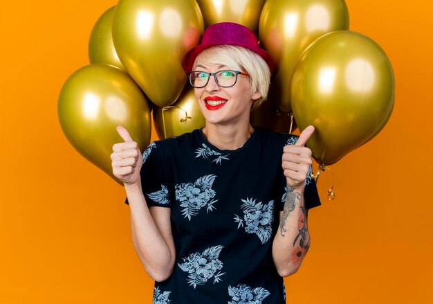 Sorridente giovane bionda festa donna che indossa il cappello del partito e occhiali in piedi davanti a palloncini guardando la parte anteriore che mostra i pollici in su isolato sulla parete arancione