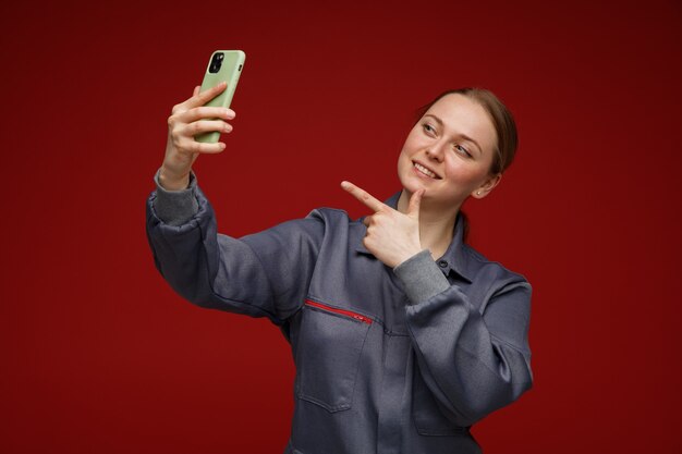 Sorridente giovane bionda ingegnere femminile che indossa uniformi tenendo selfie che punta al telefono cellulare