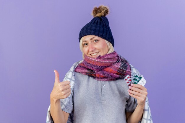 Sorridente giovane bionda malata donna slava che indossa cappello invernale e sciarpa avvolti in plaid pollici in su