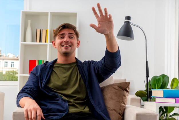 Sorridente giovane biondo bell'uomo si siede sulla poltrona alzando la mano e guardando la telecamera all'interno del soggiorno