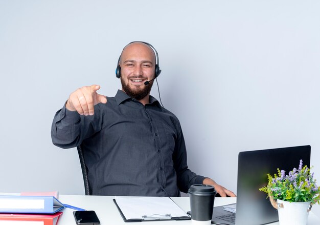 Sorridente giovane calvo call center uomo che indossa la cuffia avricolare seduto alla scrivania con strumenti di lavoro che punta davanti isolato sul muro bianco