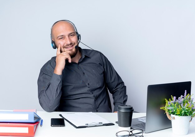 Sorridente giovane calvo call center uomo che indossa la cuffia avricolare seduto alla scrivania con strumenti di lavoro mettendo il dito sulla guancia isolato sul muro bianco