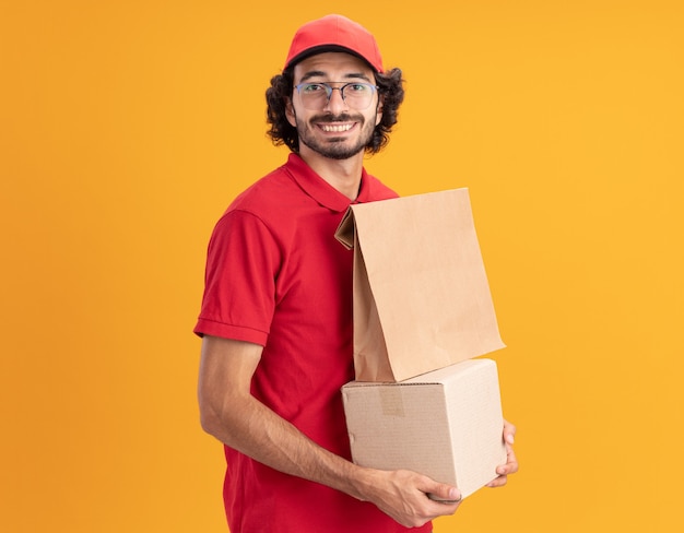 Sorridente giovane caucasica fattorino in uniforme rossa e cappello con gli occhiali in piedi in vista di profilo tenendo la scatola di cartone con il pacchetto di carta su di esso guardando davanti