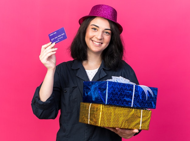 Sorridente giovane caucasica festaiola che indossa cappello da festa con pacchi regalo e carta di credito isolata sulla parete rosa