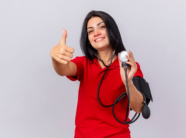 Sorridente giovane caucasica ragazza malata che indossa uno stetoscopio guardando la fotocamera misurando la sua pressione con sfigmomanometro che mostra il pollice in alto isolato su sfondo bianco con spazio di copia