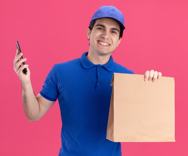 Sorridente giovane caucasico fattorino in uniforme blu e cappuccio con pacchetto di carta e telefono cellulare isolato su parete rosa on