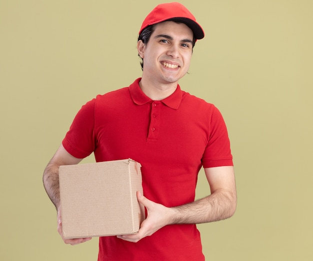 Sorridente giovane caucasico fattorino in uniforme rossa e cappuccio che tiene scatola di cartone isolato su parete verde oliva