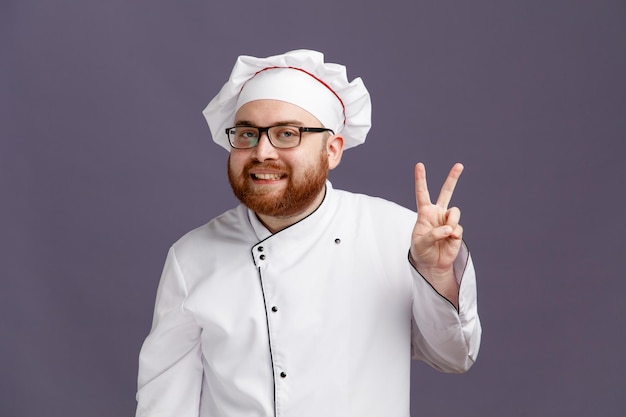 Sorridente giovane chef indossando occhiali uniforme e cappuccio guardando la fotocamera che mostra il segno di pace isolato su sfondo viola