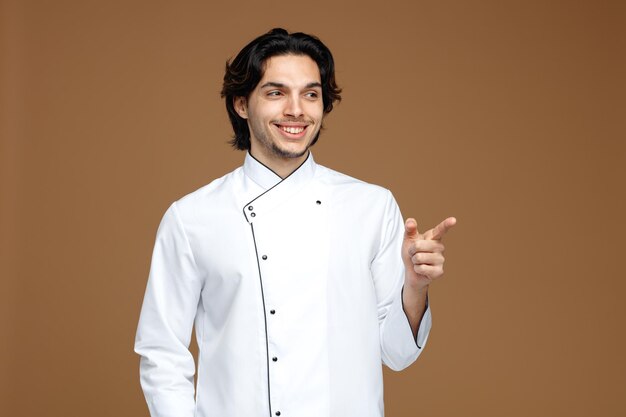 sorridente giovane chef maschio che indossa l'uniforme guardando e puntando a lato isolato su sfondo marrone