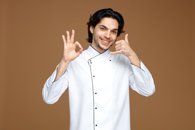 sorridente giovane chef maschio che indossa l'uniforme guardando la fotocamera che mostra segno ok e gesto di chiamata isolato su sfondo marrone