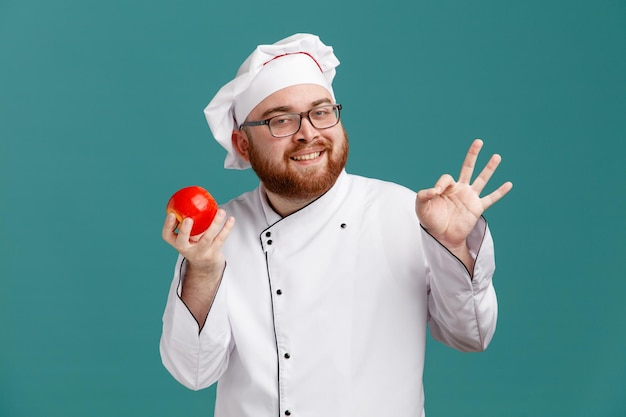 Sorridente giovane chef maschio che indossa occhiali uniforme e cappuccio che tiene mela guardando la fotocamera che mostra segno ok isolato su sfondo blu