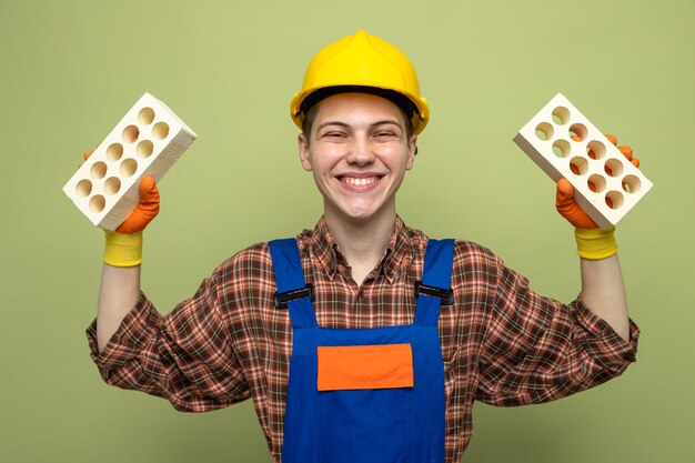 Sorridente giovane costruttore maschio che indossa l'uniforme con i guanti che tengono i mattoni