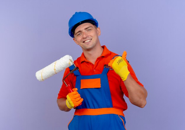 Sorridente giovane costruttore maschio che indossa l'uniforme e il casco di sicurezza in guanti che tengono il rullo di vernice il suo pollice in su sulla porpora