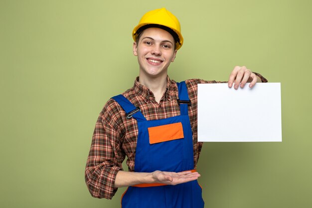 Sorridente giovane costruttore maschio che tiene e indica con la carta a mano che indossa l'uniforme