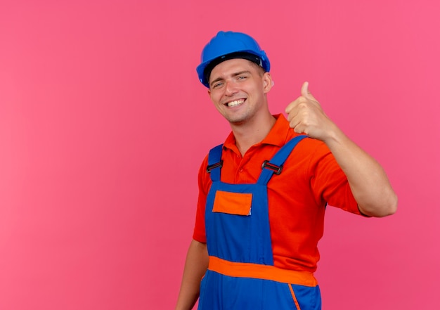 Sorridente giovane costruttore maschio indossa uniforme e casco di sicurezza il pollice in alto sul rosa