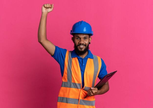 sorridente giovane costruttore uomo in uniforme con casco di sicurezza in piedi con il pugno alzato e tenendo appunti isolati sulla parete rosa con copia spazio