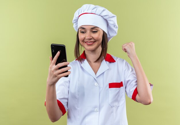 Sorridente giovane cuoca che indossa l'uniforme dello chef tenendo e guardando il telefono che mostra sì gesto isolato su parete verde oliva