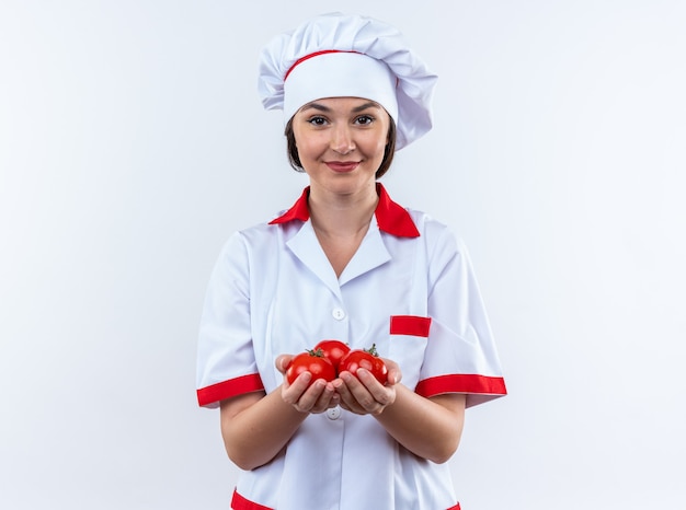 Sorridente giovane cuoca che indossa l'uniforme dello chef tenendo fuori il pomodoro in telecamera isolata su sfondo bianco