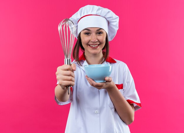 Sorridente giovane cuoca che indossa l'uniforme dello chef tenendo fuori la ciotola con la frusta in telecamera isolata su sfondo rosa