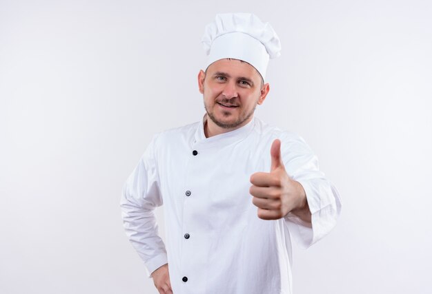 Sorridente giovane cuoco bello in uniforme del cuoco unico che mostra pollice su isolato su spazio bianco