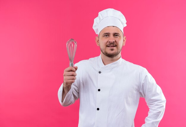 Sorridente giovane cuoco bello in uniforme del cuoco unico che tiene frusta isolato su spazio rosa