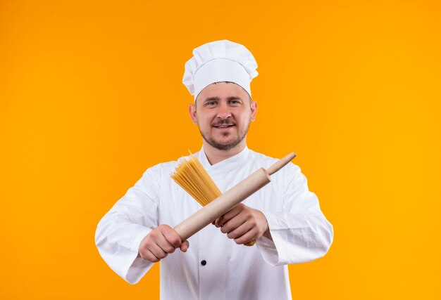 Sorridente giovane cuoco bello in uniforme del cuoco unico che tiene gli spaghetti e il mattarello isolato sullo spazio arancione