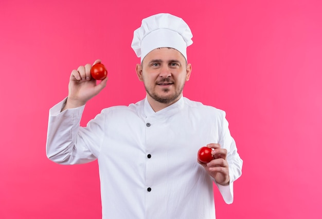 Sorridente giovane cuoco bello in uniforme del cuoco unico che tiene i pomodori isolati sullo spazio rosa