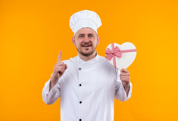 Sorridente giovane cuoco bello in uniforme del cuoco unico che tiene il contenitore di regalo a forma di cuore e che alza il dito sullo spazio arancione isolato