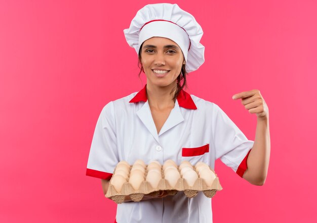 Sorridente giovane cuoco femminile che indossa la tenuta uniforme del cuoco unico e punti al lotto di uova con lo spazio della copia