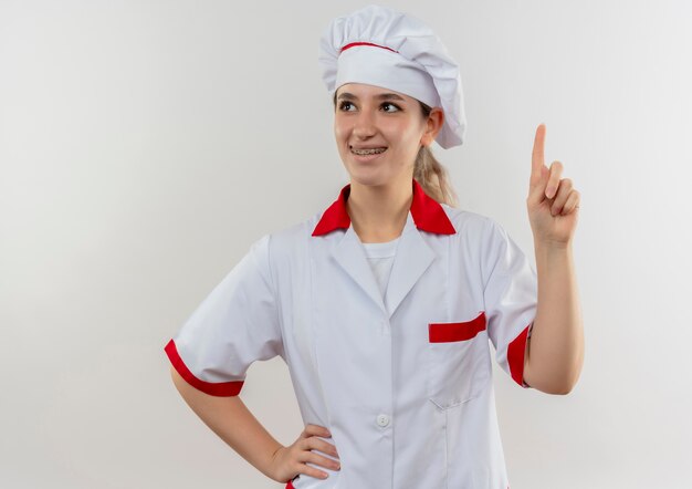 Sorridente giovane cuoco grazioso in uniforme da chef con bretelle dentali alzando il dito mettendo la mano sulla vita e guardando il lato isolato su uno spazio bianco