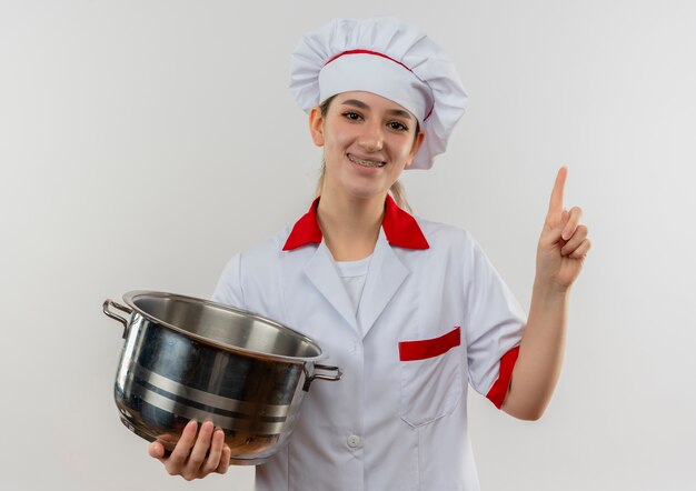Sorridente giovane cuoco grazioso in uniforme da chef con bretelle dentali che tengono pentola e alzando il dito isolato su spazio bianco