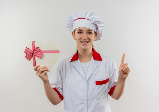 Sorridente giovane cuoco grazioso in uniforme da chef con parentesi graffe dentali che tiene il contenitore di regalo a forma di cuore e che alza il dito isolato su spazio bianco