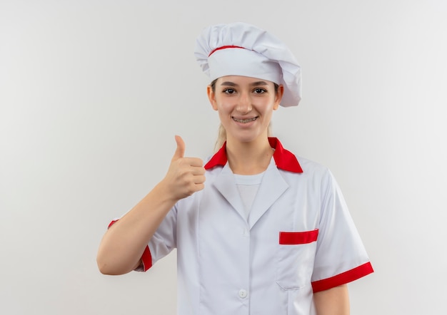 Sorridente giovane cuoco grazioso in uniforme del cuoco unico con le parentesi graffe dentali che mostra il pollice in su che sembra isolato su spazio bianco