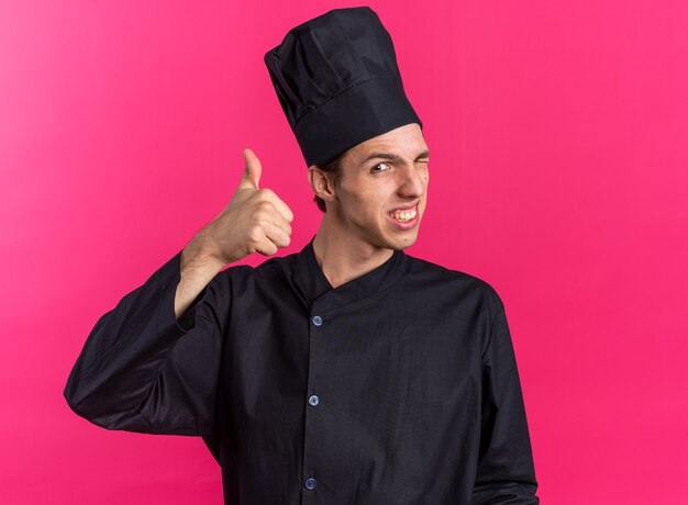 Sorridente giovane cuoco maschio biondo in uniforme da chef e berretto che guarda l'obbiettivo che fa l'occhiolino che mostra il pollice in su isolato sulla parete rosa